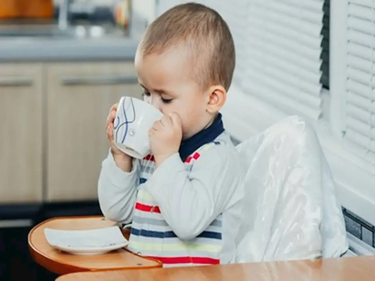 Małe dziecko siedzi w krzesełku i pije z kubeczka. Zastanawiasz się, do kiedy dziecko powinno pić mleko modyfikowane?