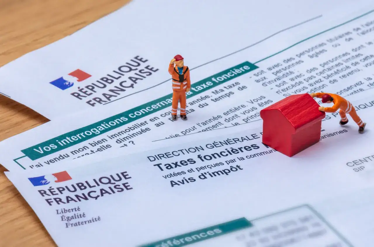 Figurines sur des documents de la R&eacute;publique Fran&ccedil;aise. L'une pousse une maison rouge, l'autre salue. Th&egrave;me : taxe fonci&egrave;re date.