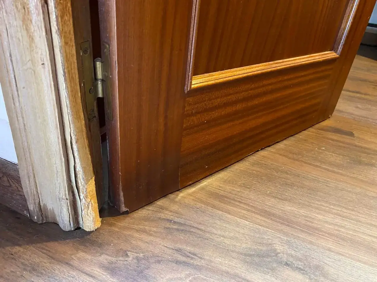 Marco de puerta de madera hinchada por humedad, con bisagra visible y suelo de madera.