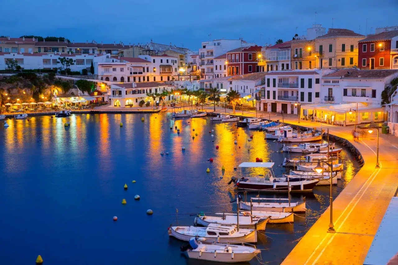 Porto Mahon illuminato di sera, con barche ormeggiate e riflessi dorati sull'acqua.