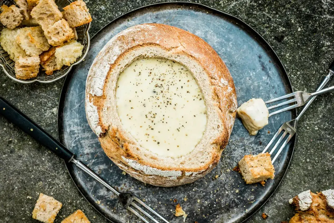 Un pain rond creusé sert de bol pour une fondue au fromage crémeuse, parsemée de poivre. Des morceaux de pain et des fourchettes sont prêts pour la dégustation.