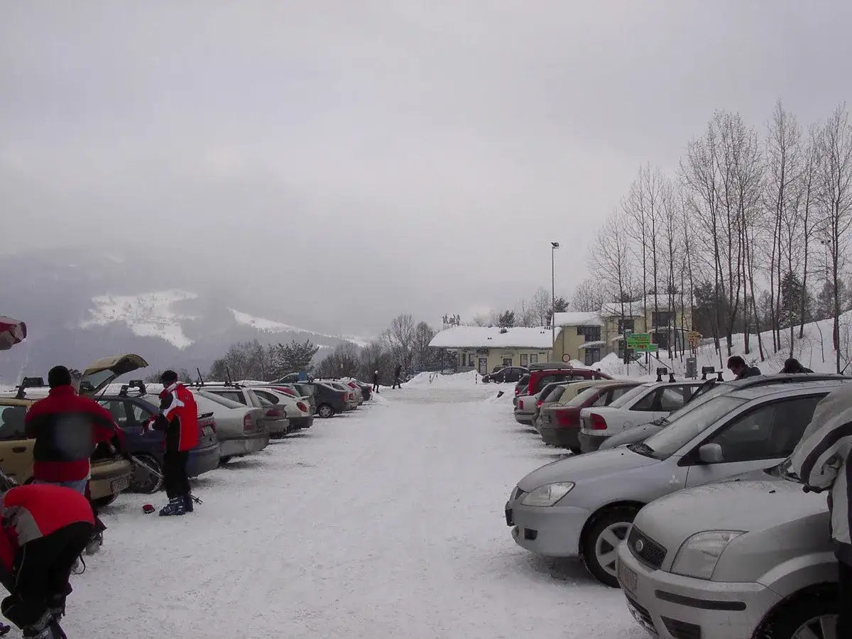 Zimowy parking pod górą Żar. Samochody stoją na ośnieżonym placu, w tle widać budynki i zaśnieżone zbocza.