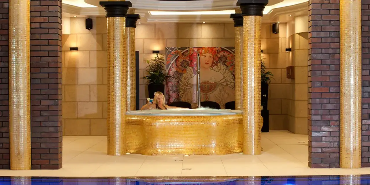 Kobieta w jacuzzi z drinkiem, otoczona złotymi mozaikami. Kolor fioletowy znaczenie &ndash; symbolizuje luksus i relaks w tym eleganckim spa.
