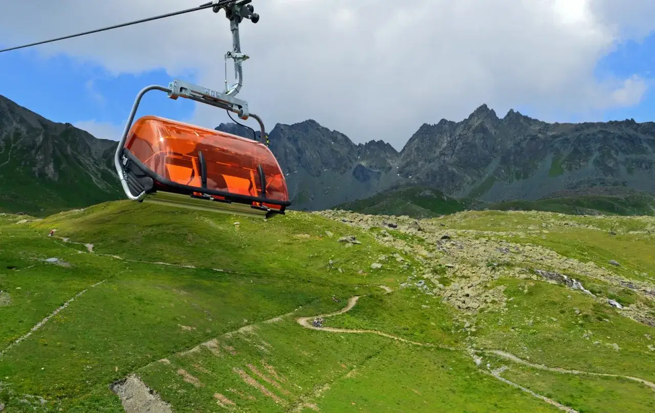 Pomarańczowa gondola kolejki linowej unosi się nad zielonymi zboczami gór w Szwajcarii. W oddali widać skaliste szczyty.