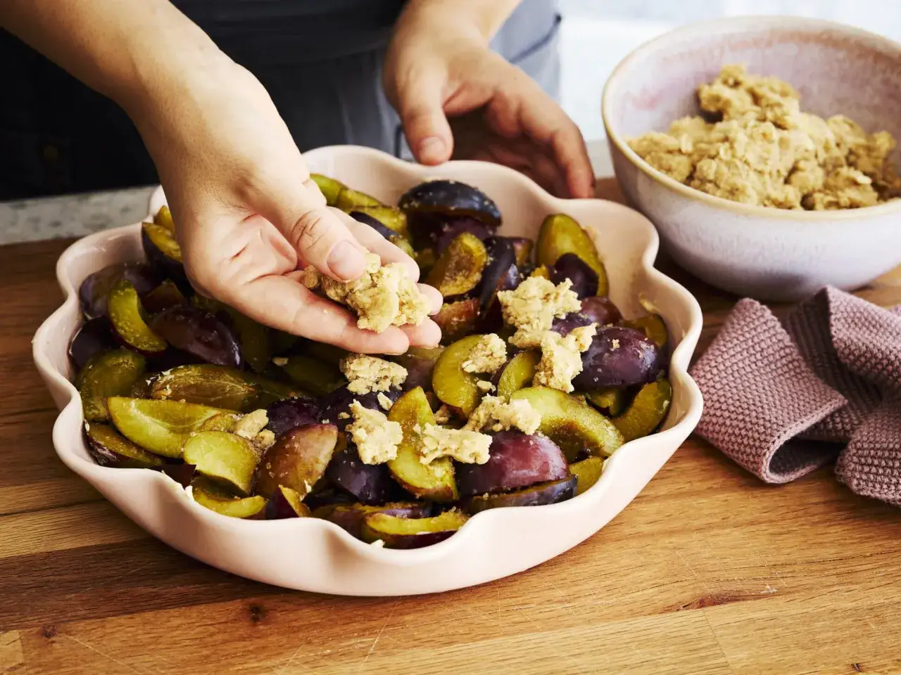 Hände verteilen Streusel auf Zwetschgen in einer rosa Form. So schnell ist ein Zwetschgen Crumble fertig!