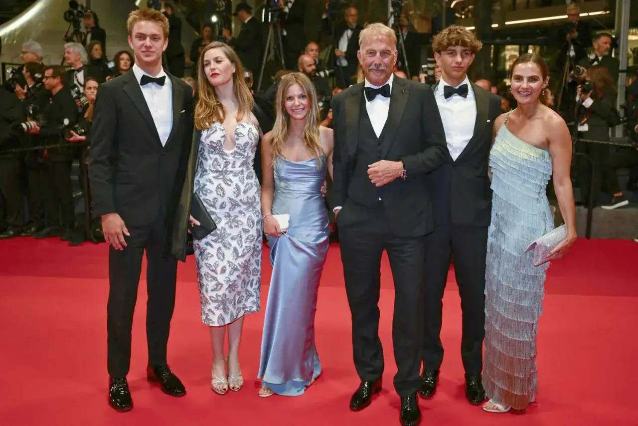 Kevin Costner z dziećmi na czerwonym dywanie. Aktor w czarnym smokingu, obok niego jego dzieci w eleganckich kreacjach.