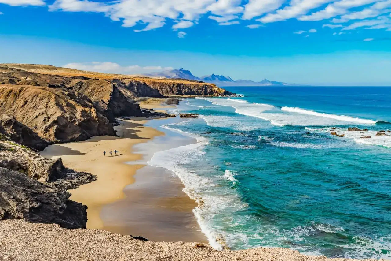 Eine malerische spanische Atlantikbucht mit felsigen Klippen, goldenem Sand und t&uuml;rkisfarbenem Wasser. Drei Surfer laufen am Strand entlang.