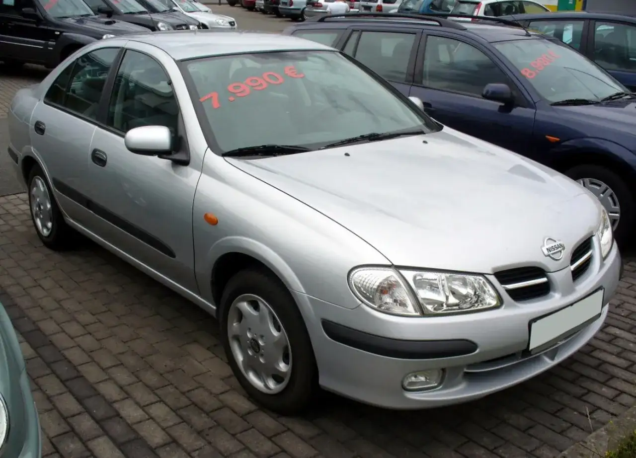Srebrny Nissan Almera z ceną 7.990€ na szybie, zaparkowany na bruku.