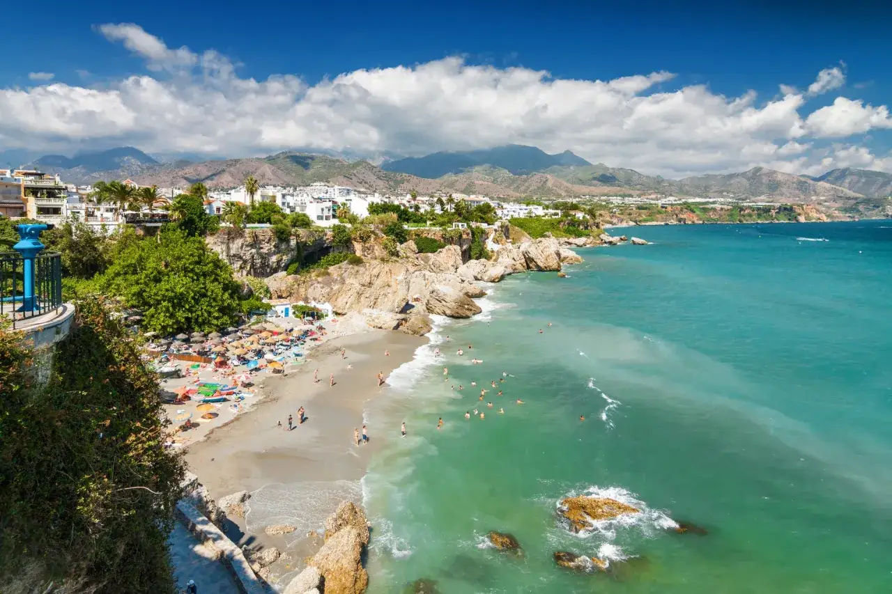 En livlig malaga strand med turkost vatten, klippor och vita hus i bakgrunden. M&auml;nniskor solar och badar.