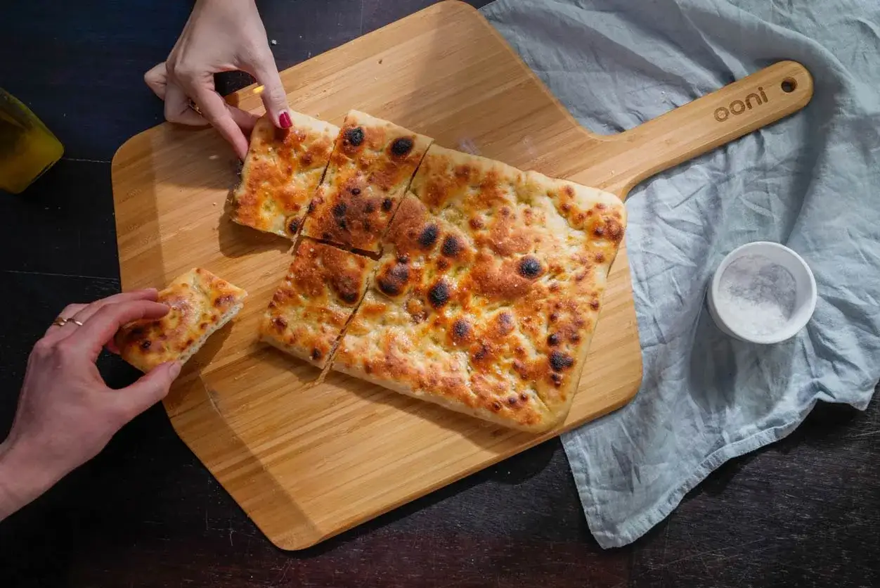 Une pizza al taglio dor&eacute;e et croustillante, coup&eacute;e en carr&eacute;s, est servie sur une planche en bois. Des mains attrapent des morceaux.
