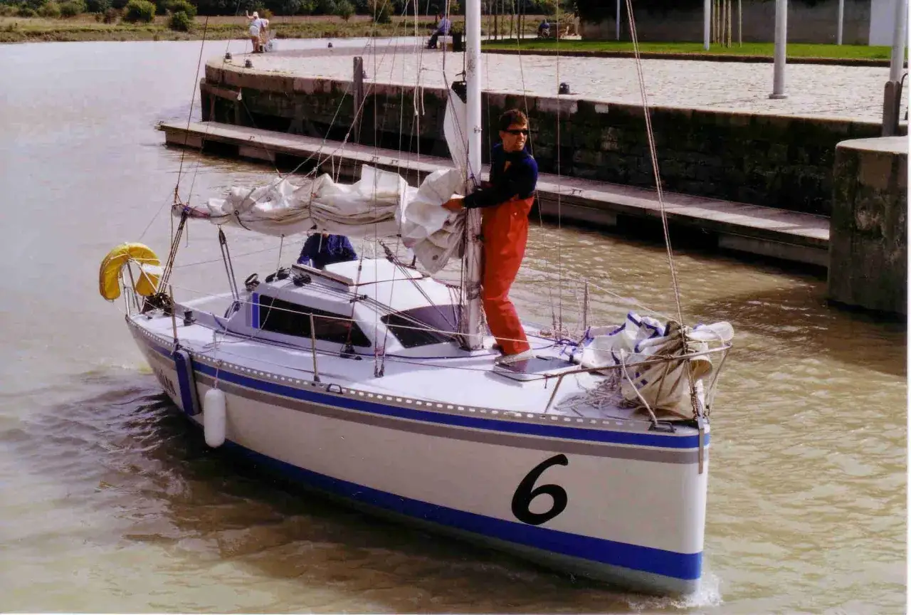 Un homme en salopette rouge prépare un voilier coco, le numéro 6 bien visible sur la proue, prêt à naviguer sur un canal.