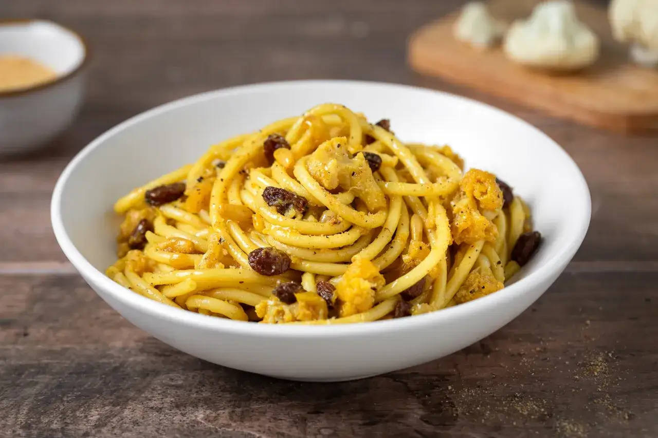 Un piatto di pasta e cavolfiore con uvetta e pangrattato, servito in una ciotola bianca su un tavolo di legno scuro.