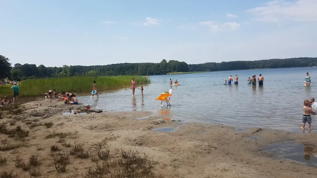 Ludzie kąpią się i relaksują na piaszczystej plaży nad jeziorem Świętajno. Woda jest spokojna, a na horyzoncie widać zalesione brzegi.