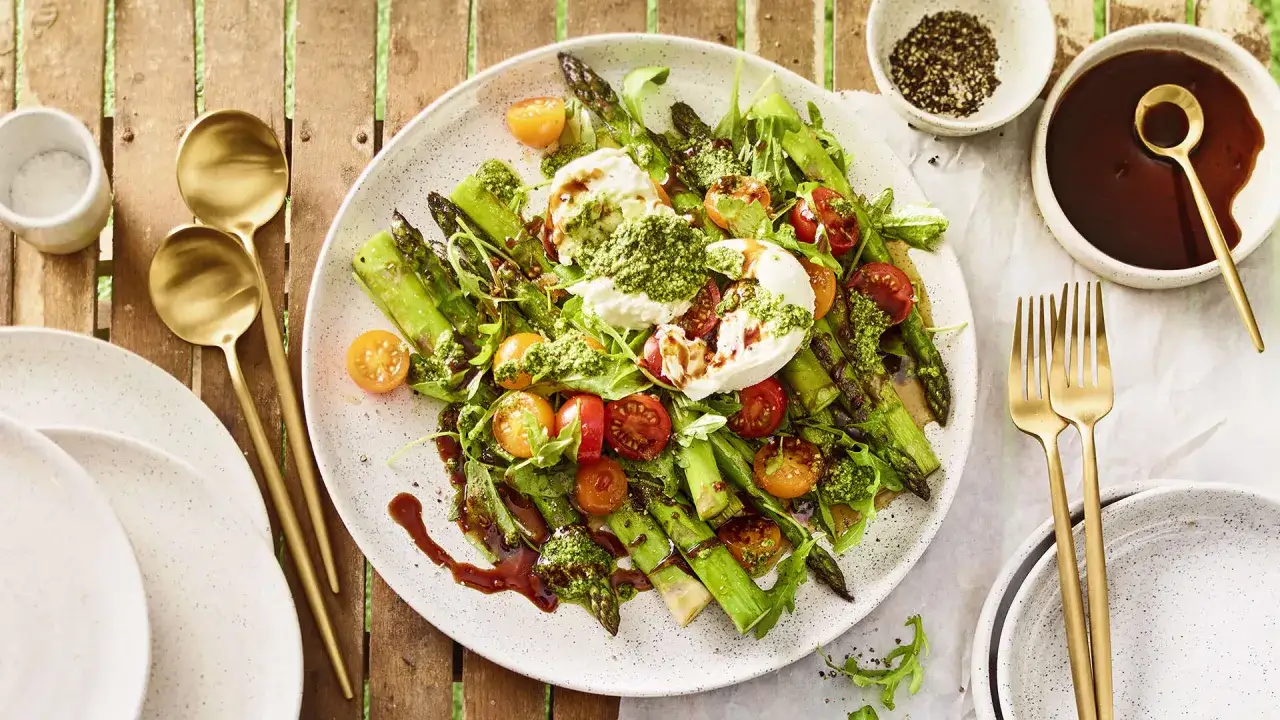 Spargel mit Burrata, Kirschtomaten und Pesto auf einem Teller, garniert mit Balsamico-Glasur. Goldene Gabeln und Löffel liegen daneben.