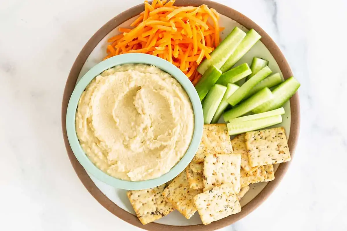En skål med krämig hummus utan tahini, serverad med gurkstavar, strimlade morötter och kex. Perfekt för ett lätt mellanmål.