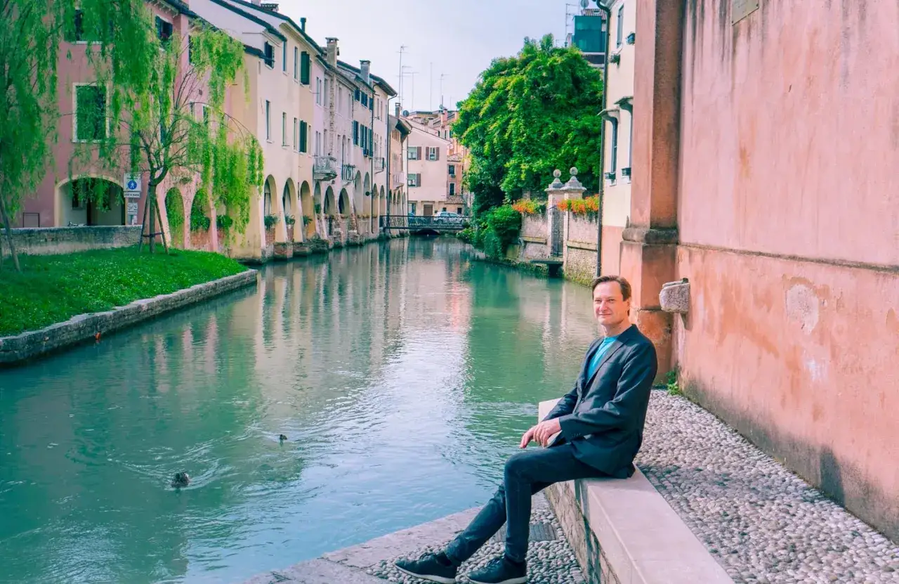 Man sitter vid en kanal i Treviso, Italien, med f&auml;rgglada hus och tr&auml;d l&auml;ngs vattnet.