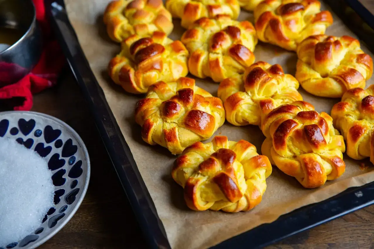 Nybakade zeina saffransbullar på en plåt, gyllene och inbjudande. Bredvid en skål med socker och en röd duk.
