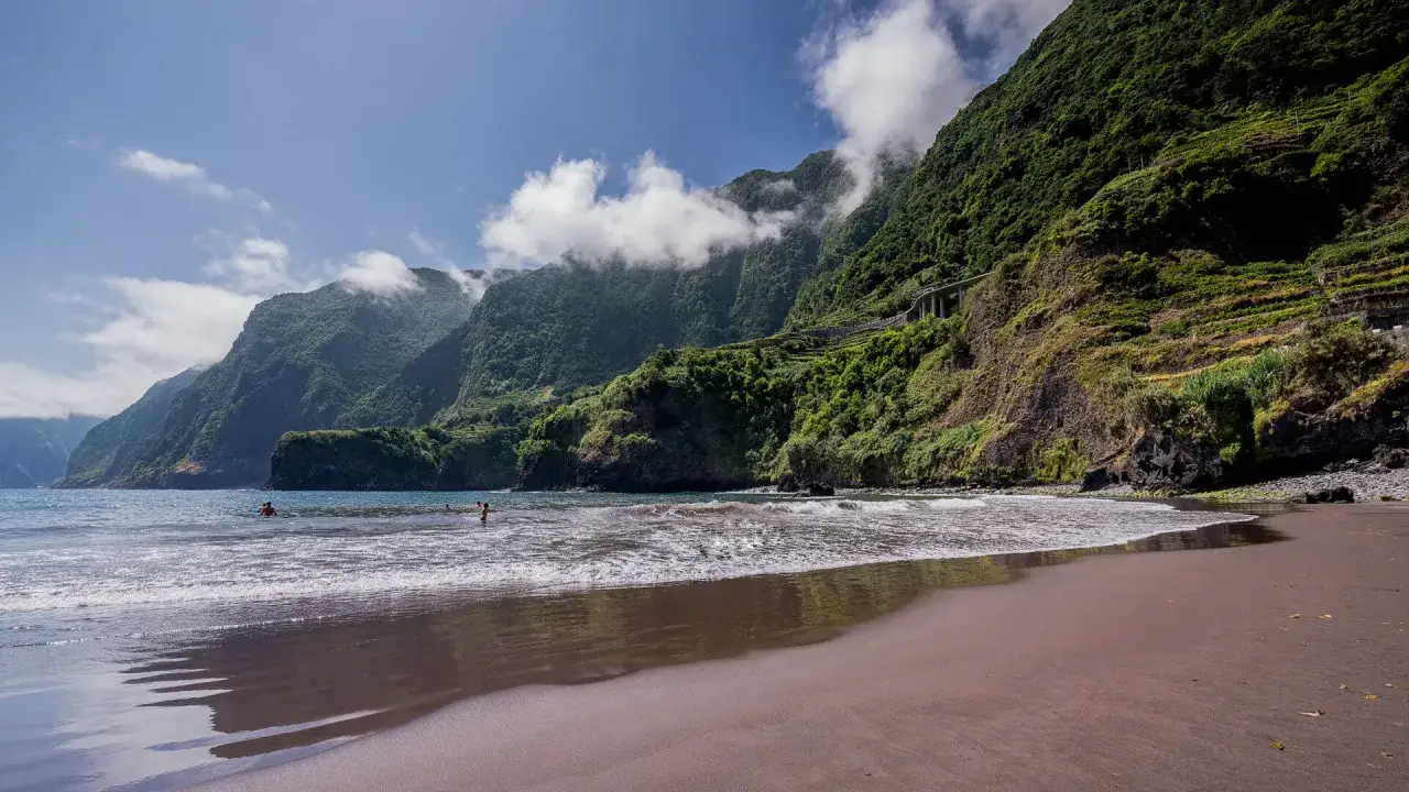 Ein malerischer Madeira Strand mit türkisfarbenem Wasser und üppig grünen Bergen im Hintergrund. Einige Menschen genießen das kühle Nass.