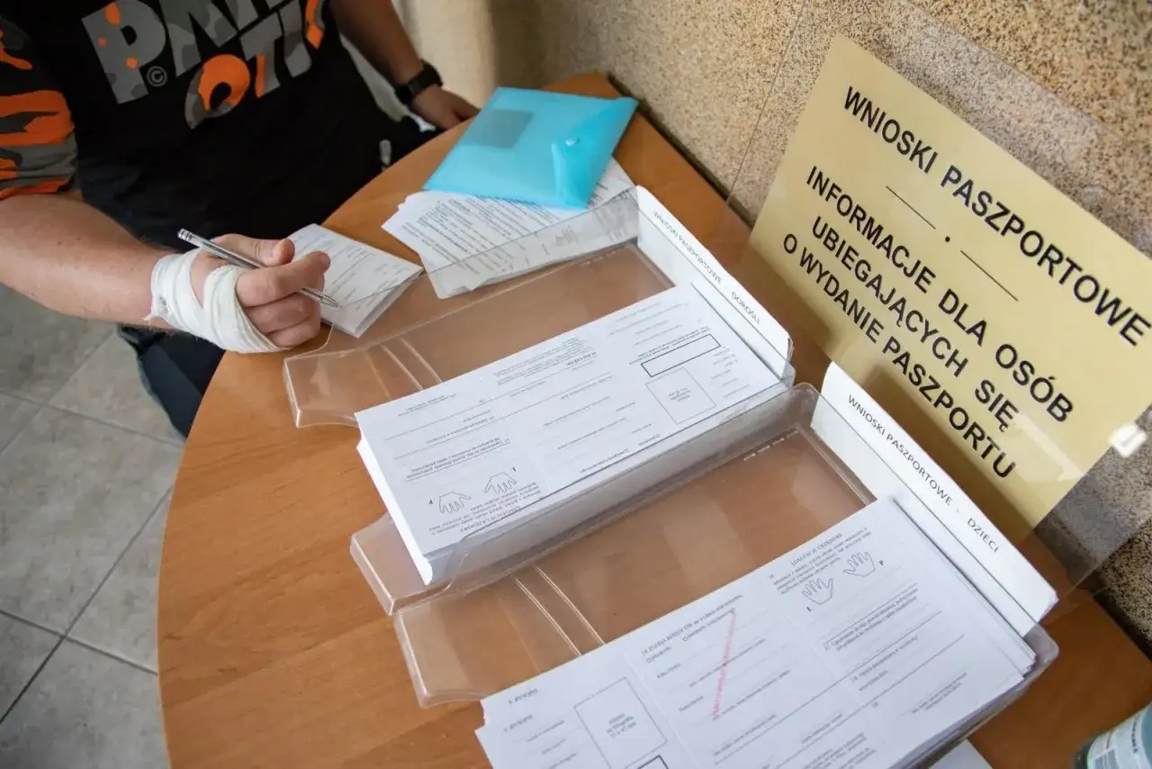 Osoba z zabandażowaną ręką wypełnia formularze, by dowiedzieć się, jak wyrobić paszport. Na stole leżą wnioski paszportowe dla dorosłych i dzieci.