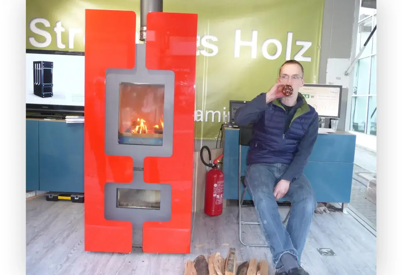 Mann trinkt vor modernem Kaminofen mit Feuer. Ein Schild im Hintergrund erwähnt
