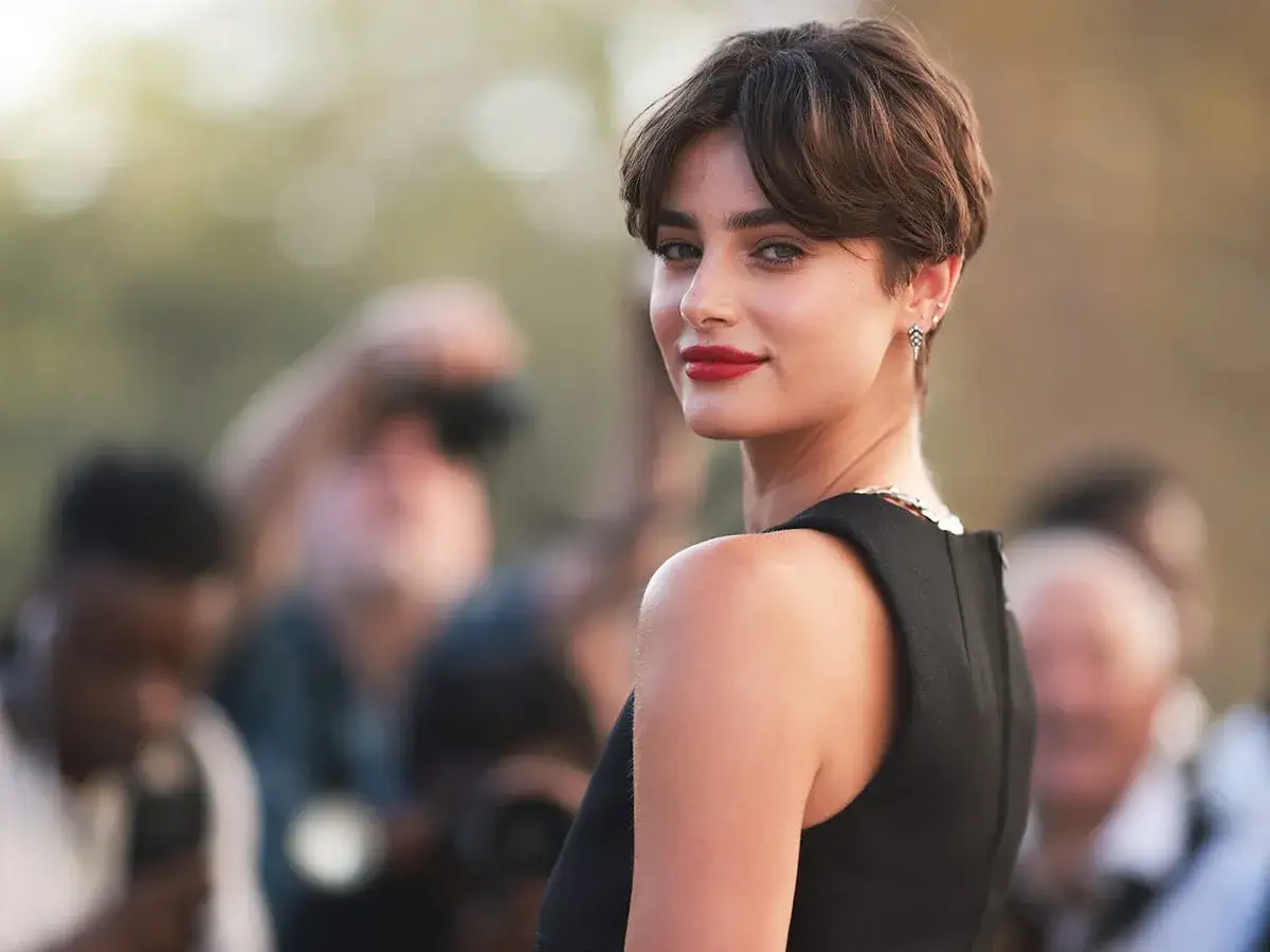 Mujer con un moderno corte bixie, labios rojos y mirada intensa, rodeada de fot&oacute;grafos.