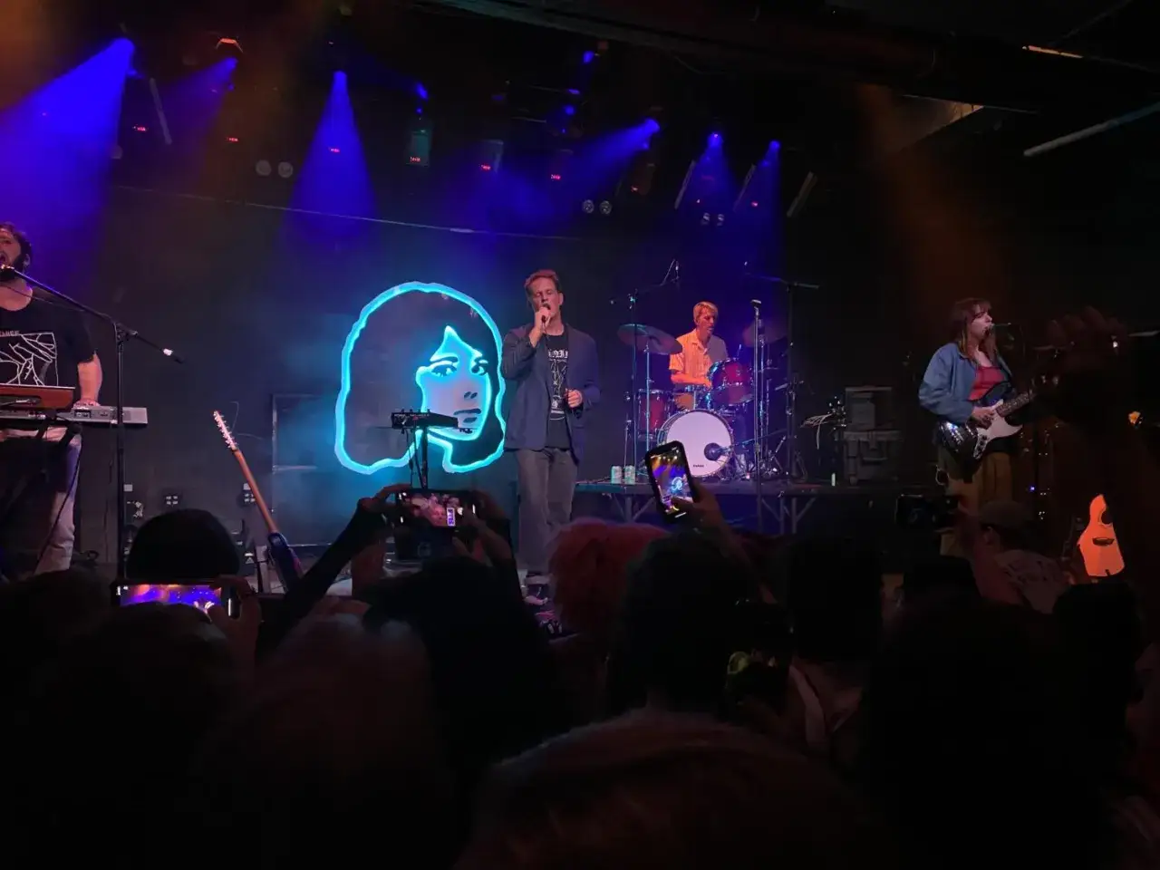 Koncert zespołu tv girl. Wokalista śpiewa na scenie, za nim neonowa podobizna dziewczyny. Publiczność filmuje telefonami.