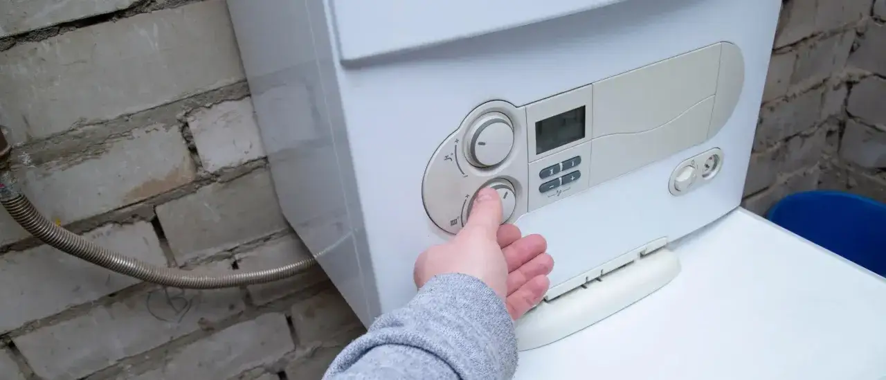 Main d'une personne ajustant le thermostat d'une chaudi&egrave;re gaz ou &eacute;lectrique, install&eacute;e contre un mur de briques.
