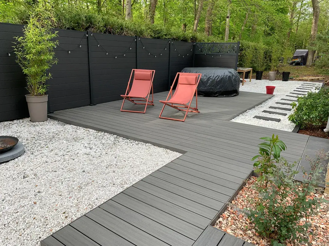 Une pose terrasse bois sur terre, avec deux chaises pliantes corail, un spa b&acirc;ch&eacute; et un chemin de dalles blanches.