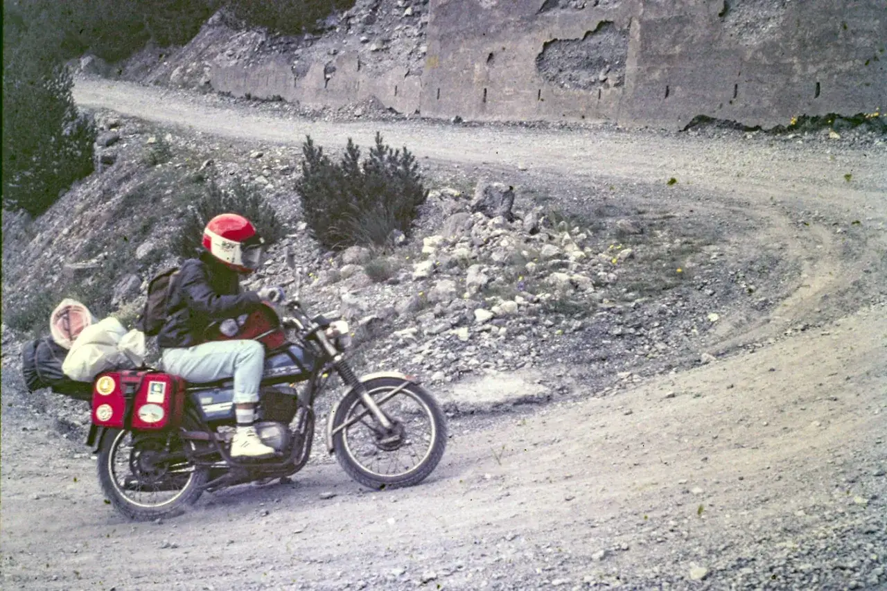 Motociclista con casco rosso affronta il passo Gavia in moto, su strada sterrata e rocciosa, con bagagli al seguito.