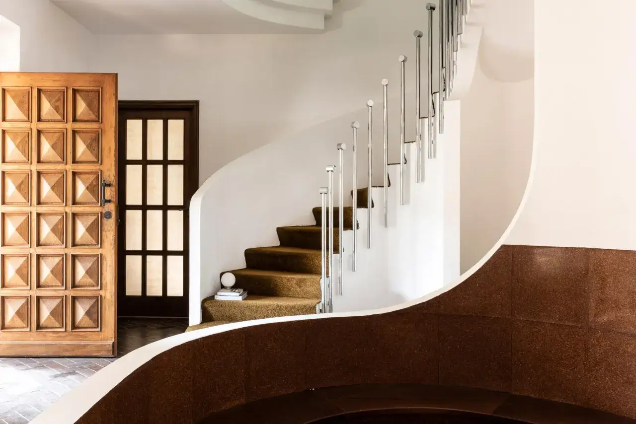 Scala interna con gradini in moquette marrone, corrimano in metallo lucido e porte in legno intagliato. Un esempio di come abbellire una scala interna con stile.