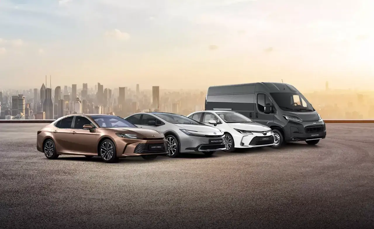 Różnorodne **toyota modele**: brązowa Camry Hybrid, srebrny Prius Plug-in Hybrid, biała Corolla i szary Proace Max na tle miasta.