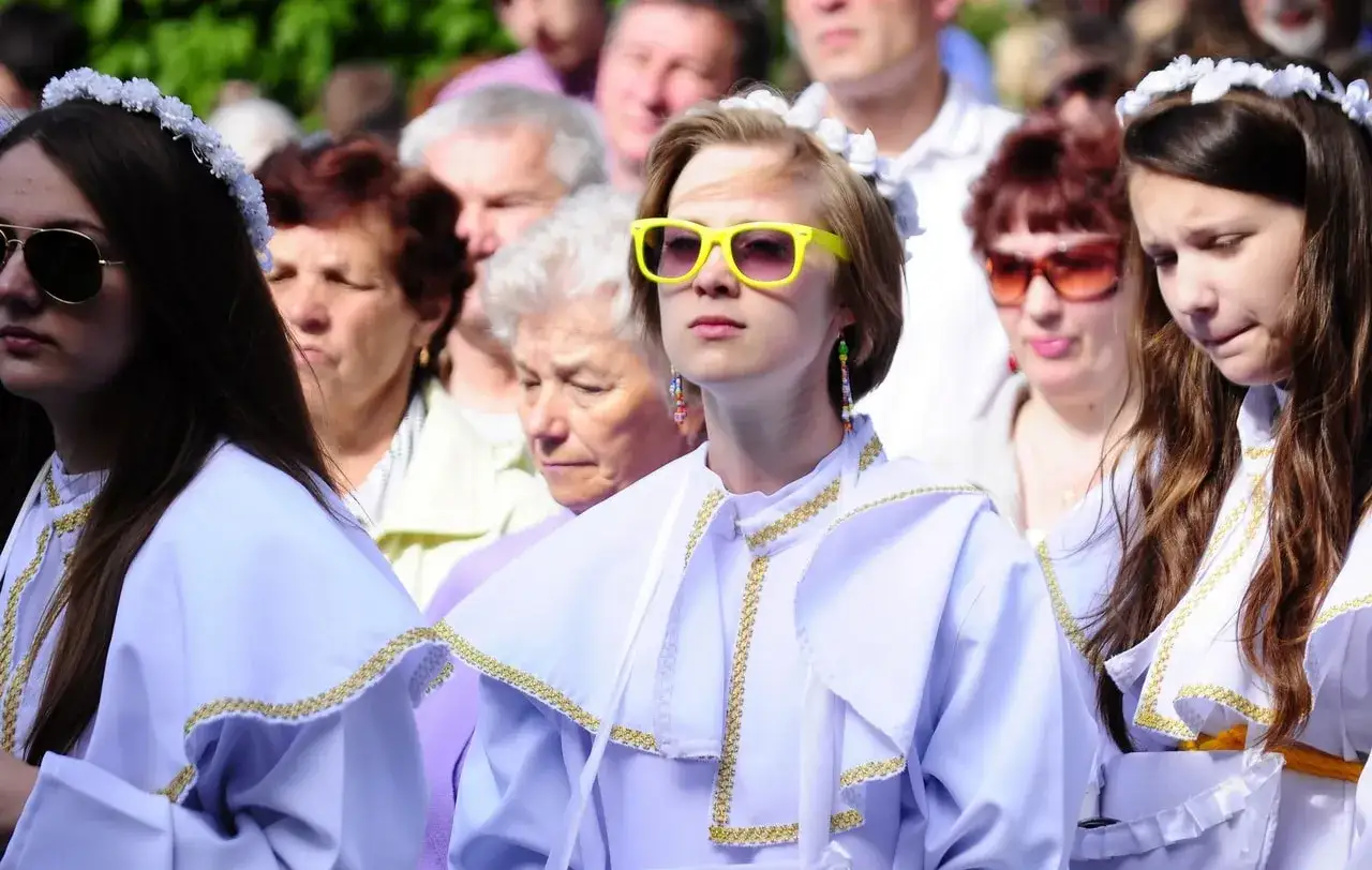 Dziewczęta w białych strojach z ozdobnymi kołnierzami, przygotowujące się do bierzmowania. Jedna z nich ma żółte okulary.