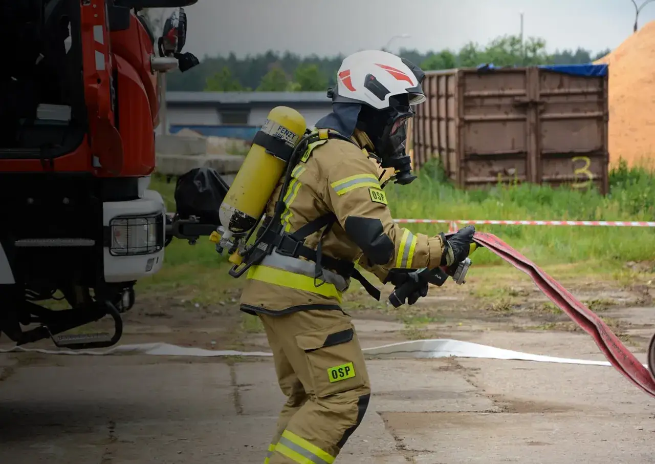 Strażak w pełnym rynsztunku, z aparatem tlenowym na plecach, przygotowuje się do akcji. Jego atrybuty strażaka – hełm, maska, rękawice i specjalistyczny wąż – świadczą o gotowości do walki z żywiołem.