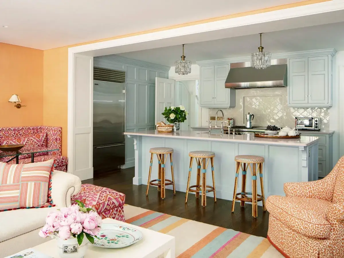 Une cuisine moderne aux armoires bleu pâle, avec un îlot central et des tabourets. La couleur mur cuisine moderne est un orange vif dans le salon adjacent.