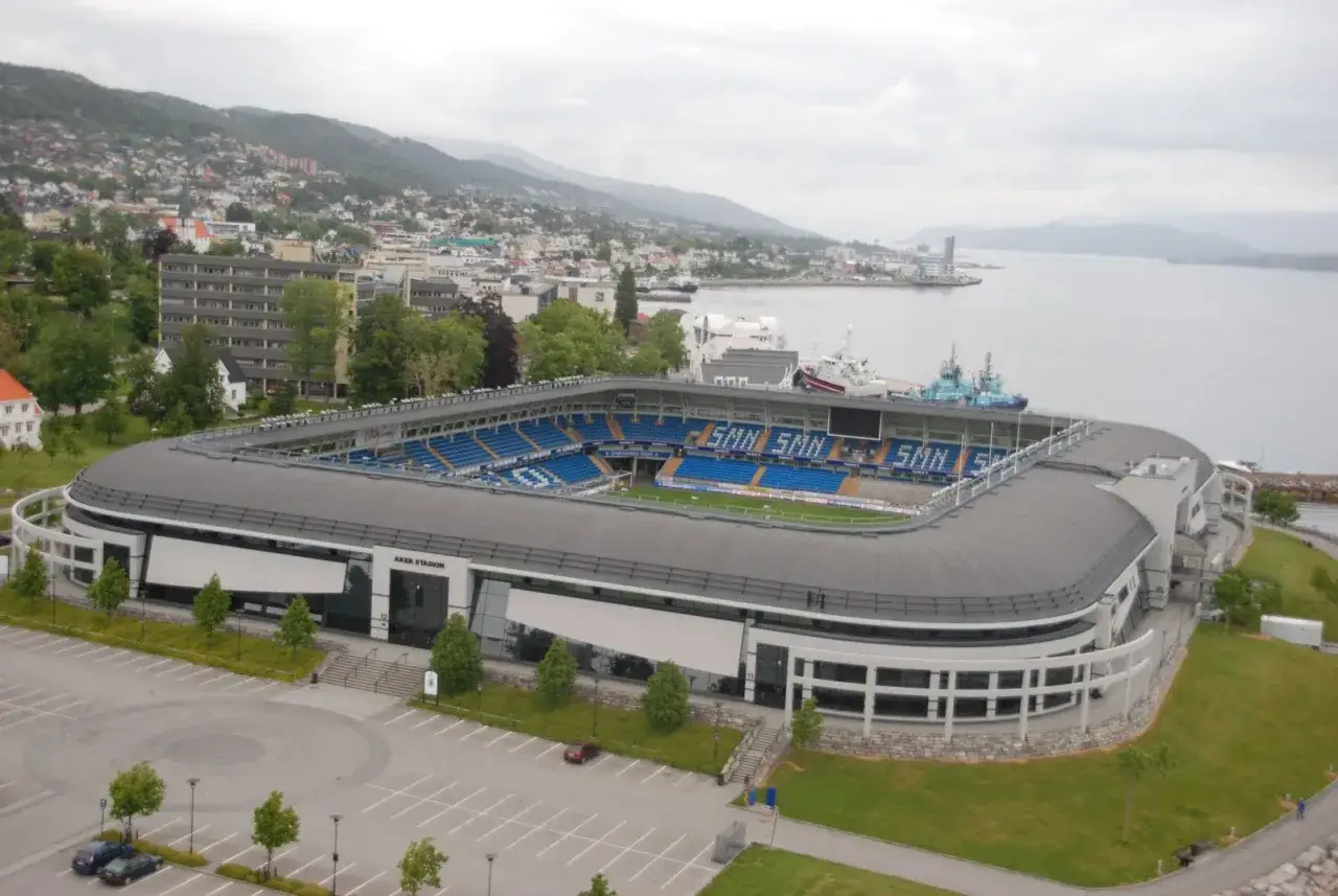 Stadion Aker w Molde, z niebieskimi trybunami i zieloną murawą, otoczony wodą i wzgórzami.