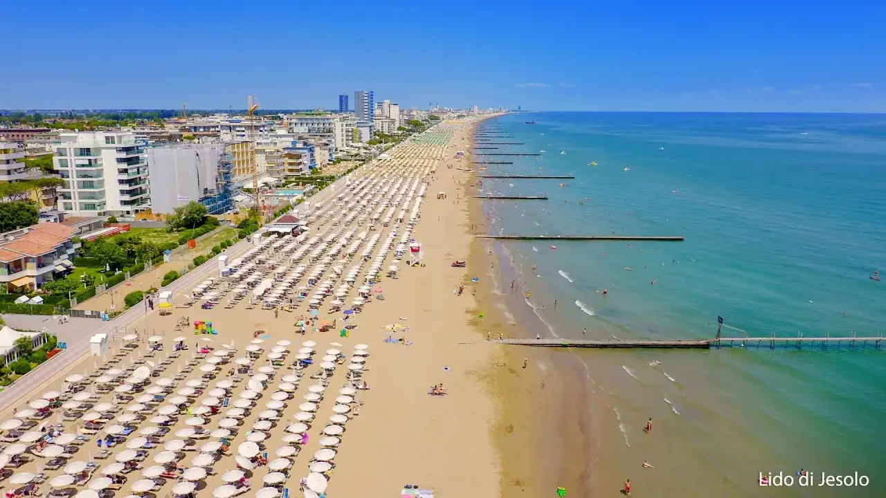 Słoneczna plaża w Lido di Jesolo z rzędami leżak&oacute;w i parasoli, turkusowe morze, molo i budynki w tle. Atrakcje dla każdego!