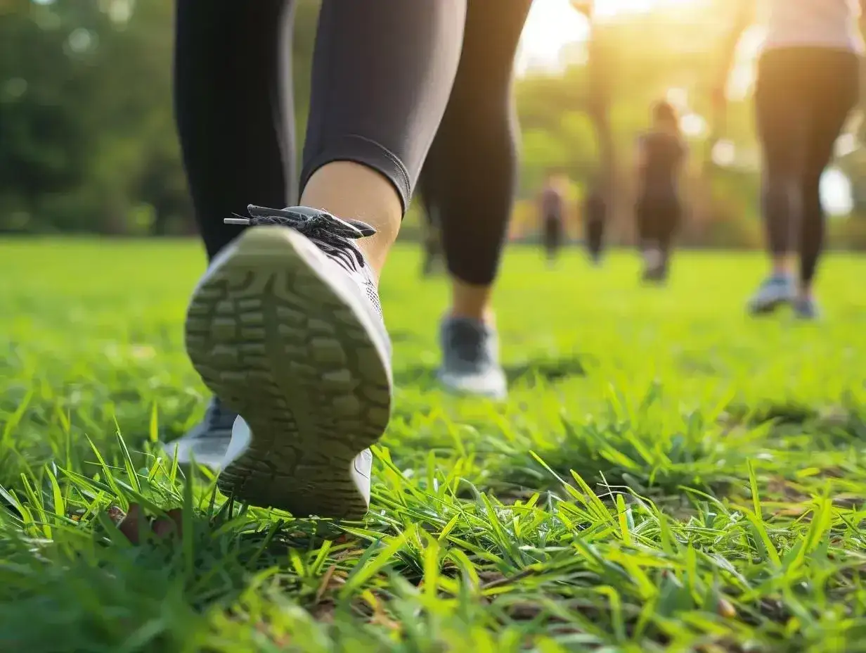 Grupa osób uprawia power walking po zielonej trawie w parku. Słońce przebija się przez drzewa.