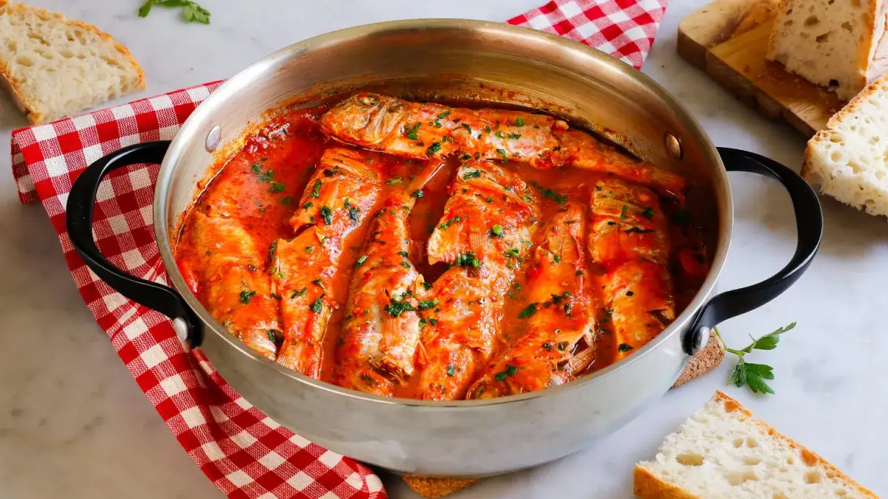 Triglie alla livornese in un tegame, con pomodoro e prezzemolo, accompagnate da fette di pane rustico.