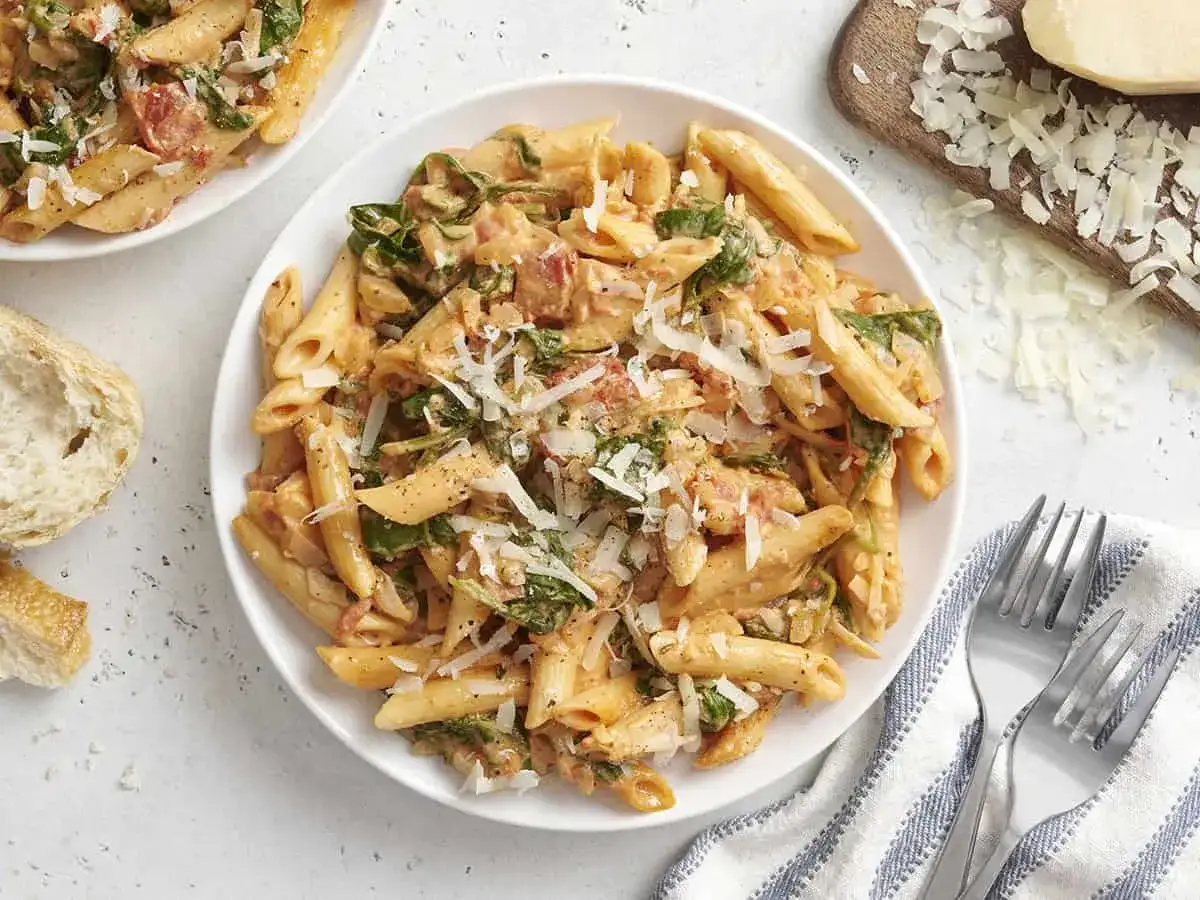 Penne w kremowym sosie z pomidorami i szpinakiem, posypane parmezanem. Obok kromka chleba i widelce.