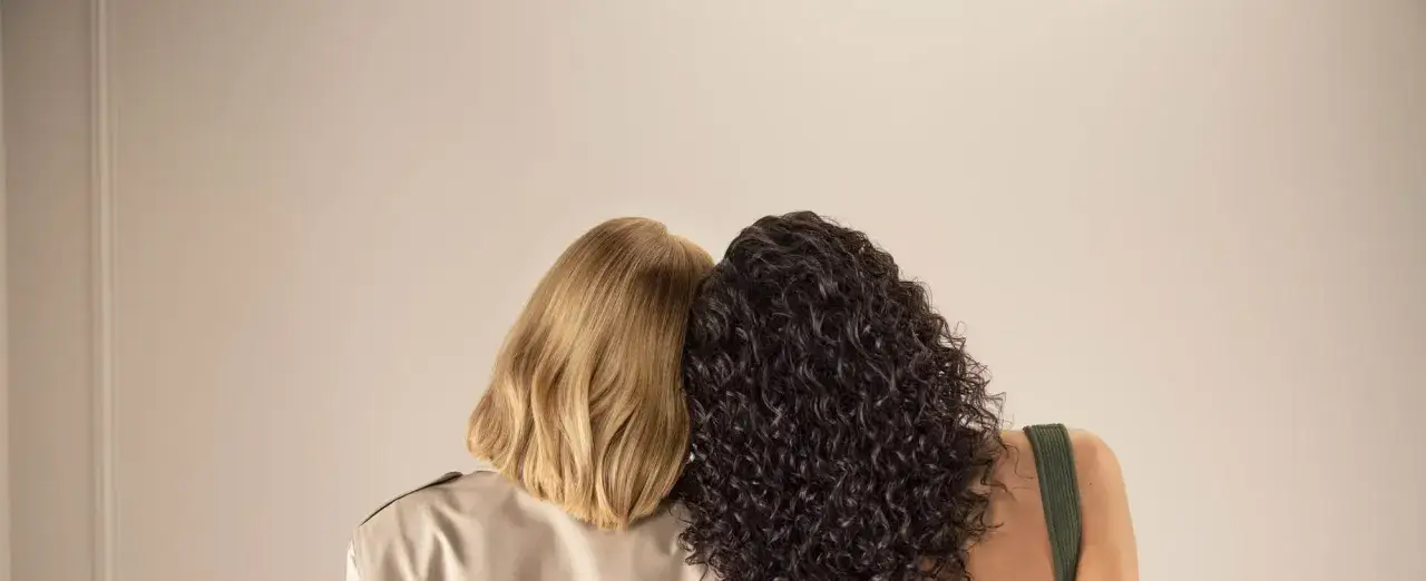 Due donne, una con capelli biondi lisci e l'altra con capelli scuri ricci, si abbracciano. La domanda è quanto dura una tinta permanente.
