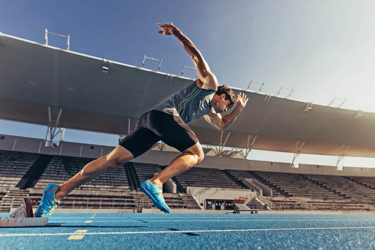 Sprinter w pozycji startowej, gotowy do rozpoczęcia treningu szybkościowego na niebieskiej bieżni stadionu.