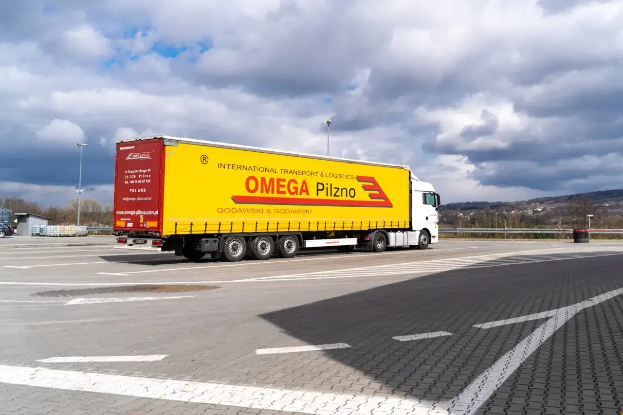 Biały tir z żółtą naczepą OMEGA Pilzno na parkingu. Zastanawiasz się, ile pali tir z naczepą? Ten pojazd pokonuje długie trasy.