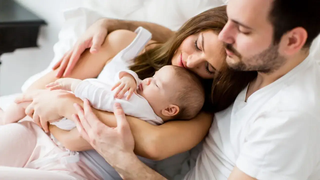 Gl&uuml;ckliche Familie im Bett. Mutter und Vater halten ihr Baby, das friedlich schl&auml;ft. Ein Moment des Sonderurlaubs nach der Geburt.