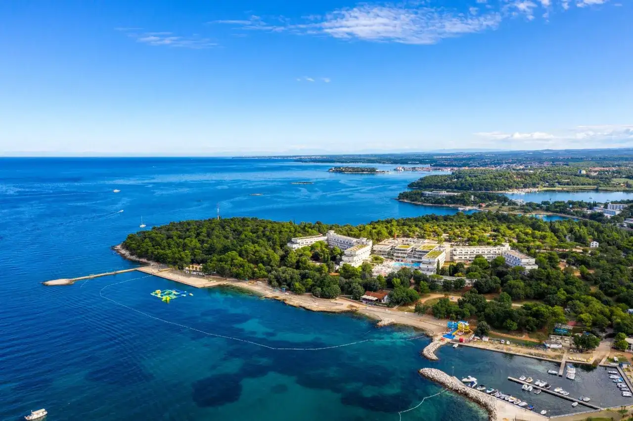 Hotel Delfin Plava Laguna z lotu ptaka, otoczony zielenią i błękitnym morzem. Widok na plażę, baseny i przystań z łodziami.