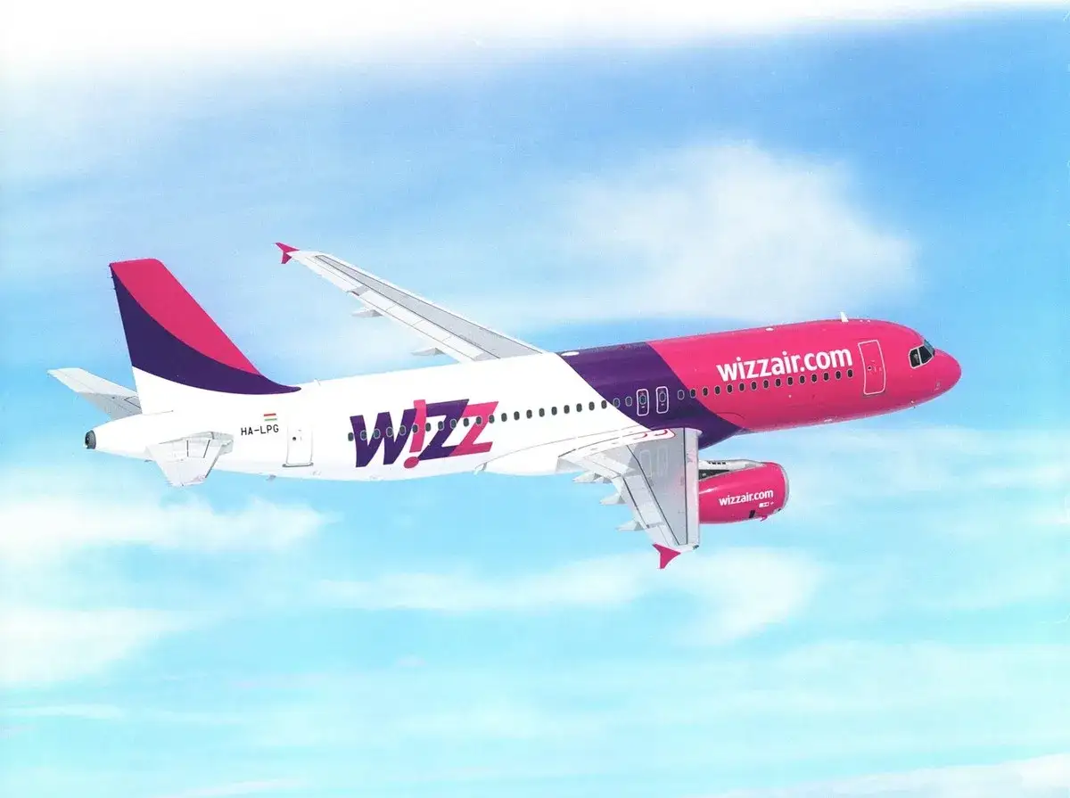Samolot Wizz Air w różowo-fioletowych barwach leci na tle błękitnego nieba z chmurami. Wizz Air to znana linia lotnicza.