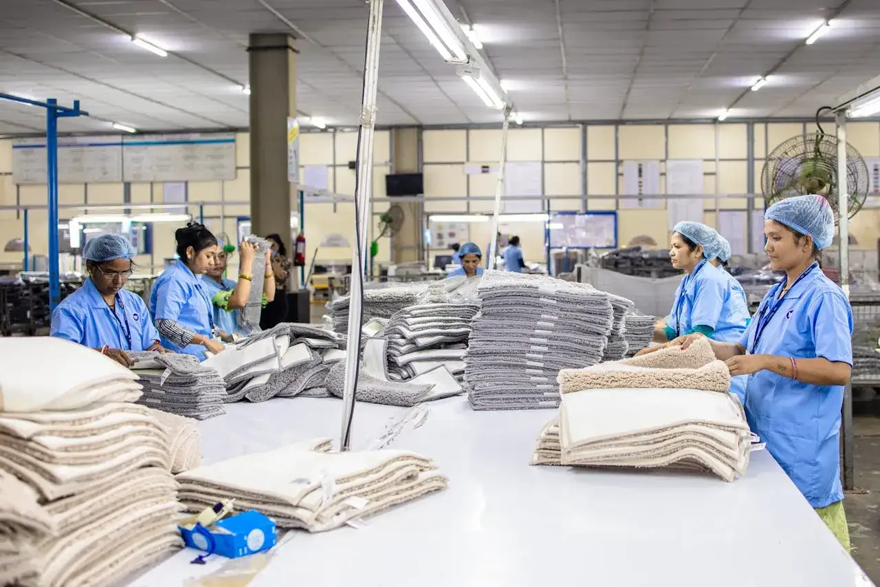 Kobiety w niebieskich uniformach i czepkach pracują na produkcji, składając stosy dywaników.