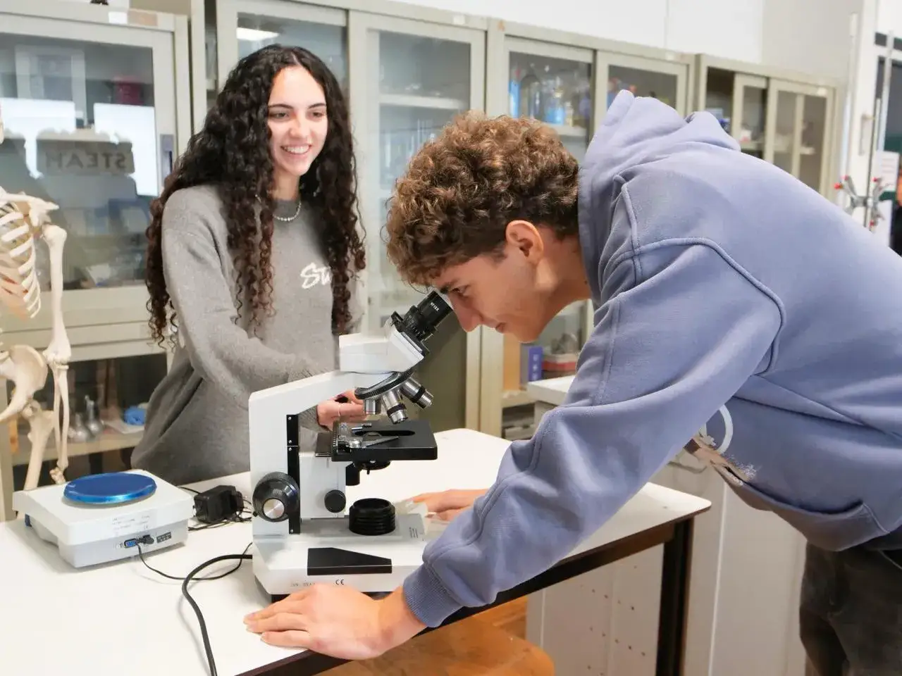 Ragazzi in laboratorio studiano scienze umane con un microscopio, bilancia e scheletro.