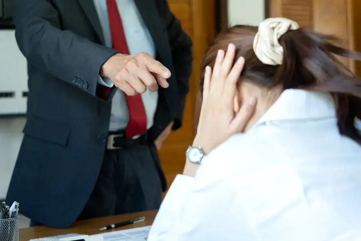 Szef wskazuje palcem na pracownika, kt&oacute;ry ukrywa twarz w dłoniach. Widać tu negatywne cechy pracownika, takie jak brak odporności na stres.