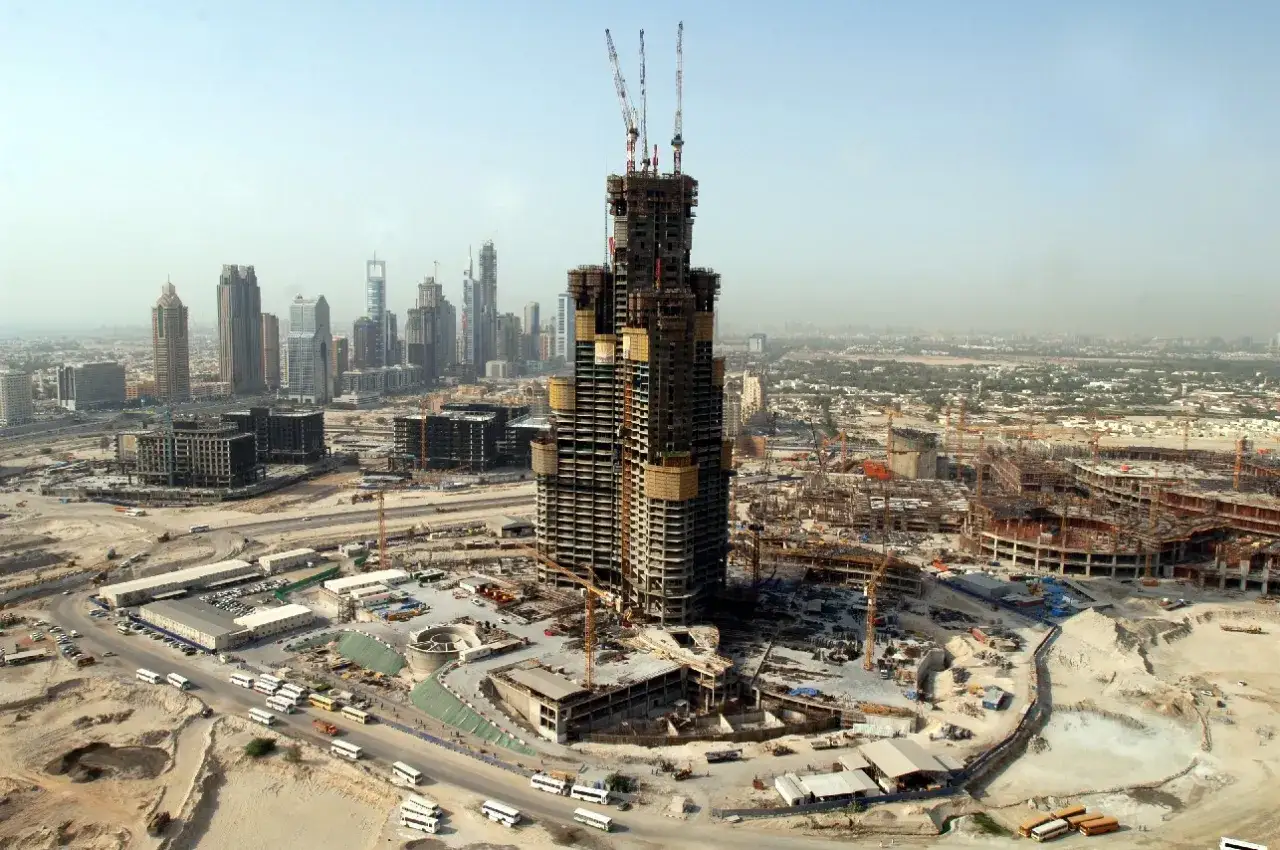 Widok na budowę Burj Khalifa, pokazujący ogromną skalę projektu i postęp prac. Widać liczne dźwigi i rusztowania, a także otaczające tereny budowy.