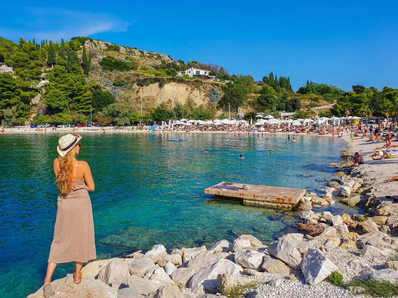 Kobieta w kapeluszu podziwia turkusowe morze i zatłoczone split plaże. Słońce, piasek i mn&oacute;stwo ludzi cieszących się wakacjami.